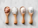 Various salt types in 4 wooden spoons on white marble.